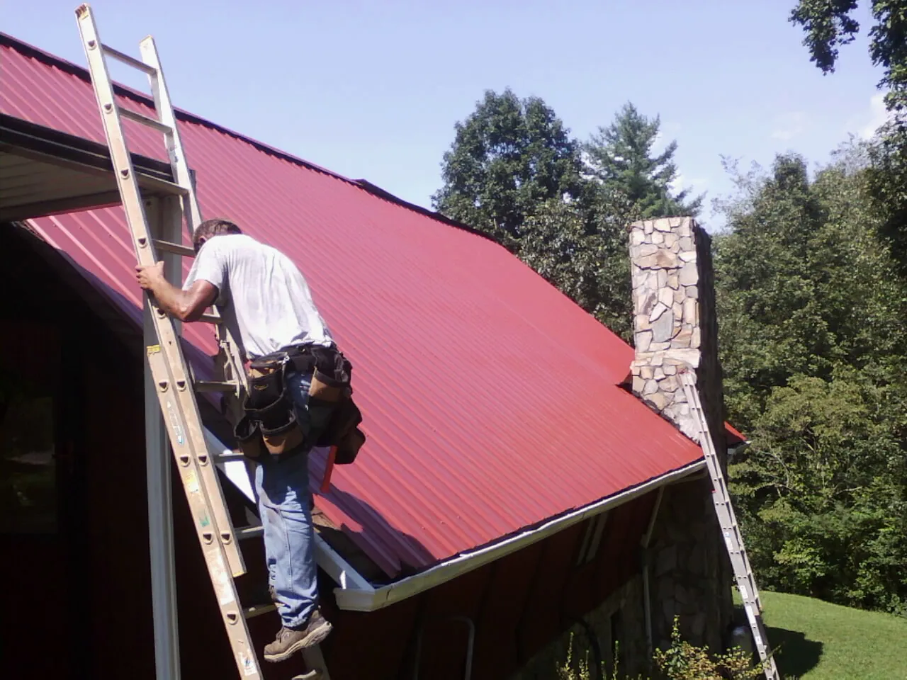 Roofer on a ladder beside a red metal roof and stone chimney — Soffit & Fascia Repair by Roofing Brothers in Flintstone